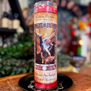 May include: A tall cylindrical red wax candle in a clear glass jar. The label features the text Madame Phoenix, Michael Archangel, and Michel Archange, with an illustration of an angel in blue robes standing over a figure in a dark setting.