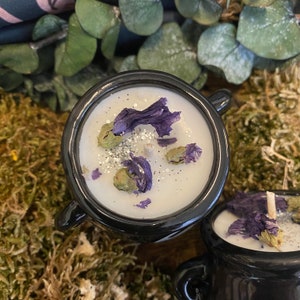 May include: Black cauldron candle with a white wax top. The wax is decorated with purple and green dried flowers and silver glitter.