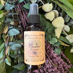 May include: A dark amber glass spray bottle with a black pump nozzle. The beige label features the text Madame Phoenix, Fluorite fluorite, and Crystal Spray, decorated with a mandala graphic. The bottle is surrounded by dried eucalyptus leaves.
