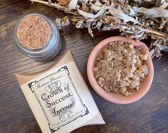 May include: A brown paper bag with the text "Madame Phoenix's Crown of Success Incense" printed on it. The bag is filled with brown incense. The incense is in a small terracotta bowl.