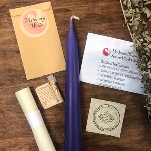May include: A purple candle, a small bottle of oil, a rolled parchment scroll, a small pouch, and a parchment square with a sigil printed on it. The items are arranged on a wooden surface. The text on the parchment square reads "Madame Phoen. Second Sight Spell Kit Contains: 1 Dipped Candle with Cotton Wick, 1 Herbal Sachet of Dressing Powder, 1 Anointing Oil Blend, 1 Parchment Talisman, 1 Parchment Scroll."