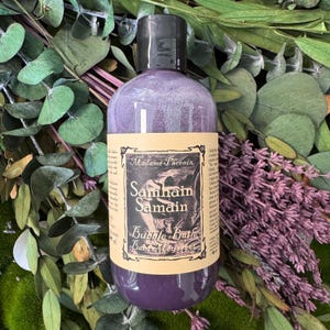 May include: A purple bubble bath bottle with a black cap, labelled "Madame Phoenix Samhain Samain Bubble Bath". The bottle is surrounded by green leaves and purple lavender sprigs, creating a natural, spa-like aesthetic.