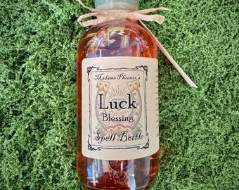 May include: A clear glass bottle filled with a reddish-brown liquid, topped with a light green cap and tied with a brown ribbon. The label reads "Luck Blessing Spell Bottle" with a horseshoe graphic. The bottle is set against a green background.