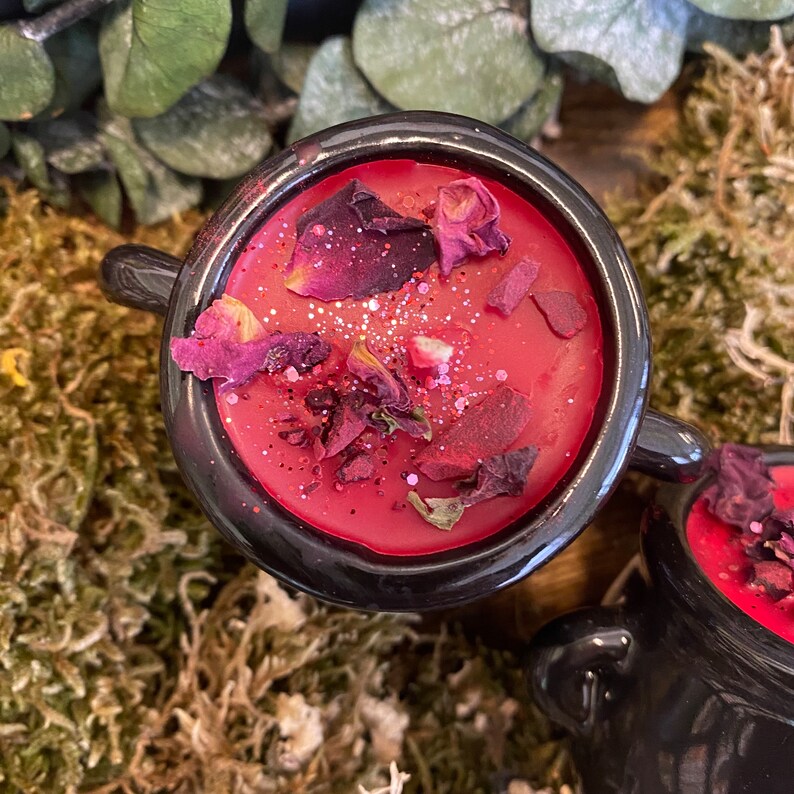 May include: A black cauldron candle with a red wax base and dried rose petals. The candle is surrounded by green moss.