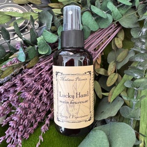 Peut inclure: Un flacon pulvérisateur brun foncé avec un bouchon noir et un embout pulvérisateur transparent. L'étiquette indique "Madame Phoenix Lucky Hand main heureuse Spray / Vaporisateur". Le flacon est entouré de lavande violette et de feuillage vert.
