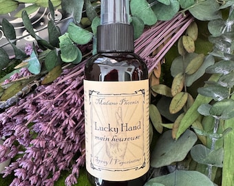 May include: A dark brown spray bottle with a black cap and a clear spray nozzle. The label reads "Madame Phoenix Lucky Hand main heureuse Spray / Vaporisateur". The bottle is surrounded by purple lavender and green foliage.