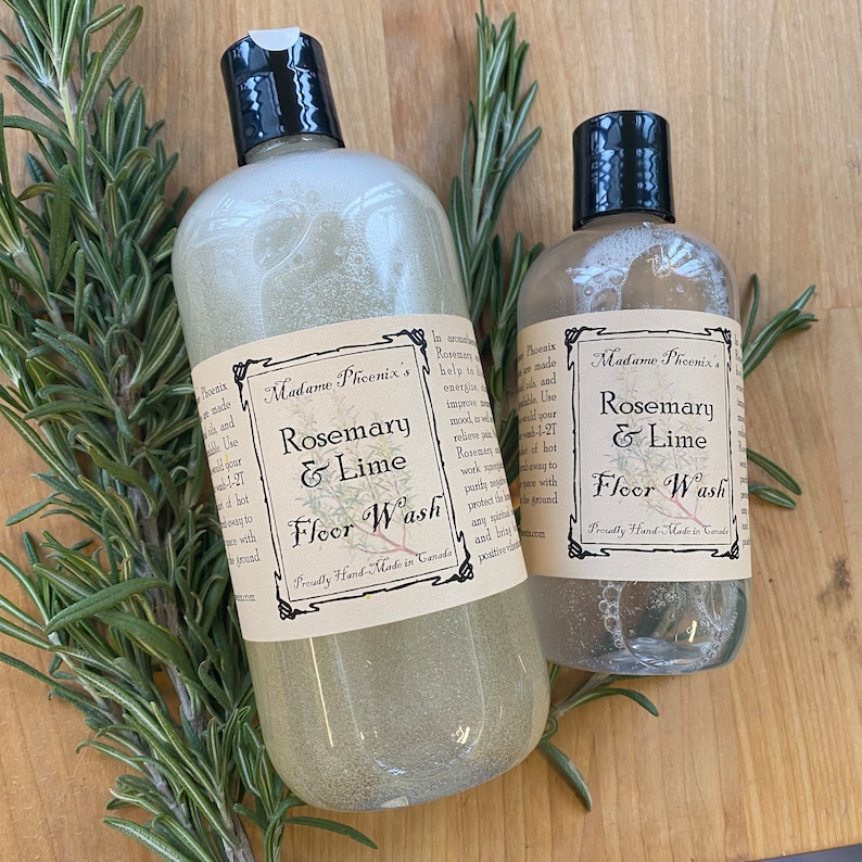 May include: Two clear bottles of Rosemary & Lime Floor Wash with black caps and labels. The labels read "Madame Phoenix's Rosemary & Lime Floor Wash". Fresh rosemary sprigs are arranged around the bottles on a wooden surface.