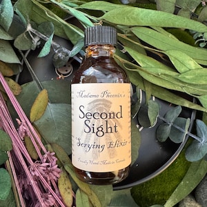 May include: A brown glass bottle with a black cap, labelled "Madame Phoenix's Second Sight Scrying Elixir." The label features an eye illustration. The bottle is surrounded by green and purple foliage.