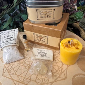 May include: A Madame Phoenix Healing Energy Spell Kit set including a metal tin, a cardboard box, a bag of bath salt, a small glass vial, a citrine crystal in a plastic bag, and a yellow pillar candle with embedded dried botanical pieces.