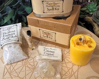 May include: A Madame Phoenix Healing Energy Spell Kit set including a metal tin, a cardboard box, a bag of bath salt, a small glass vial, a citrine crystal in a plastic bag, and a yellow pillar candle with embedded dried botanical pieces.