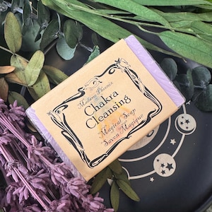 May include: A bar of lavender-colored soap in a beige paper wrapper with the words "Chakra Cleansing Magical Soap" in black and gold script. The soap is surrounded by dried lavender and greenery on a black surface.
