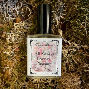 May include: A perfume bottle with a black cap and a label reading "Madame Phoenix" and "All Rites of Love & Pleasure Eau de Parfum". The label has a floral design. The bottle is on a bed of moss and dried plants.