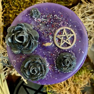May include: Purple candle with black roses and a silver pentagram charm. The candle is decorated with glitter.