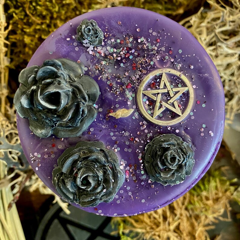 May include: Purple candle with black roses and a silver pentagram charm. The candle is decorated with glitter.