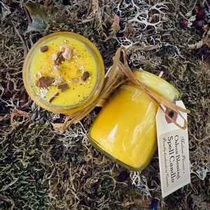 May include: A yellow candle in a glass jar with a label that reads "Oshun Blessing Spell Candle" and "Beautifully handmade in Canada". The candle is surrounded by moss.