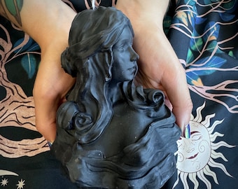 May include: A black candle shaped like a woman's bust, with long flowing hair and a detailed face. The candle is held in two hands against a dark floral background.
