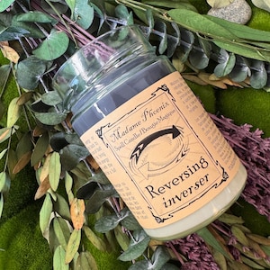 Può includere: Una candela in barattolo di vetro con cera grigio scuro e un'etichetta di carta color cuoio. L'etichetta riporta il testo Madame Phoenix, Spell Candle, Bougie Magique e Reversing inverser, insieme a un grafico a freccia circolare. Circondata da verde.