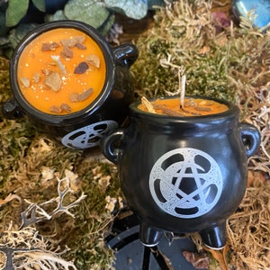 May include: Two black cauldron-shaped candles with orange wax and a pentagram design. The candles are sitting on a bed of moss.