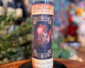 May include: A tall cylindrical glass spell candle featuring a label with the text Madame Phoenix, Queen of the Dead, and La Reine des Morts. The label displays a dark ornate frame surrounding a central illustration of a whole and sliced pomegranate.
