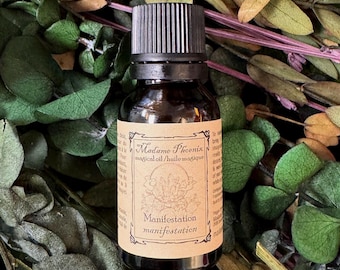 May include: A small amber glass bottle with a black ribbed screw cap and a tan paper label. The label features the text Madame Phoenix magical oil/huile magique and Manifestation manifestation with a central circular illustration of crystals.