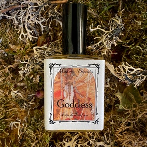 May include: A small glass bottle of perfume with a black cap. The label is white with black text and a vintage illustration of a woman with wings. The text on the label reads "Madame Phoenix Goddess Eau de Parfum".