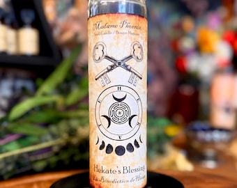 May include: A tall cylindrical glass candle filled with dark wax, featuring a beige label with the text Madame Phoenix, Spell Candle, Hekate's Blessing, and La Bénédiction de Hécate. The label displays two crossed keys, a circular labyrinth, and moon phases.
