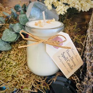 May include: A white candle in a clear glass jar with a brown twine bow and a tag that reads "House Blessing Spell Candle"  Proudly Handmade in Canada. The candle is surrounded by dried flowers and greenery.