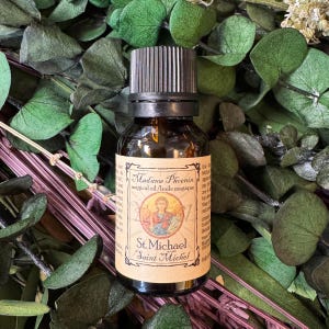 May include: A small amber glass bottle with a black ribbed cap and a tan label. The label features the text Madame Phoenix magical oil, St. Michael Saint Michel, and an illustration of a figure in robes holding a sword and scales.