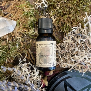 May include: A small brown glass bottle with a black cap, labelled "Madame Phoenix's Cleopatra Magical Oil Blend". The bottle is surrounded by dried moss and other natural elements.