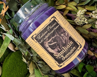 May include: A purple wax candle in a clear glass jar with a beige label. The label features the text Madame Phoenix Spell Candle/Bougie Magique and Samhain Samain, along with a dark illustration and printed descriptive text in English and French.