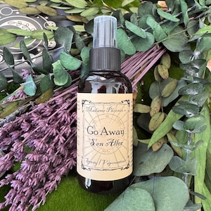 May include: A dark brown spray bottle with a black cap and a label that reads "Go Away S'en Aller Spray / Vaporisateur." The bottle is surrounded by purple lavender and green foliage.