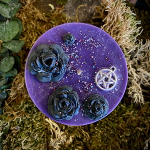 May include: Purple candle with black roses and a silver pentagram charm. The candle is decorated with glitter.