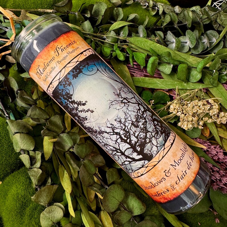 May include: A cylindrical glass candle filled with dark wax, featuring a label with the text Madame Phoenix Spell Candle and Shadows & Moonlight. The label depicts a full moon behind bare tree branches with orange and black borders.