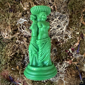May include: A green ceramic candle holder featuring two figures in a standing pose. The figures are sculpted in a classical style and are wearing flowing robes. The candle holder has a circular base and a decorative top.