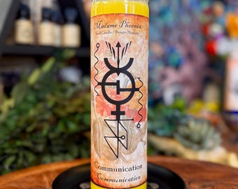 May include: A yellow pillar candle in a clear glass jar with a label featuring the text Madame Phoenix Spell Candle, Communication, and a black geometric symbol over a floral background. The candle sits on a small black circular tray.