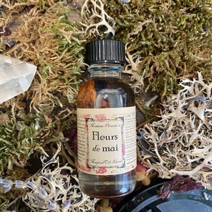 Może przedstawiać: Mała szklana butelka z czarną zakrętką wypełniona brązową cieczą. Etykieta zawiera napis "Madame Phoenix's Fleurs de mai Magical Oil Blend".