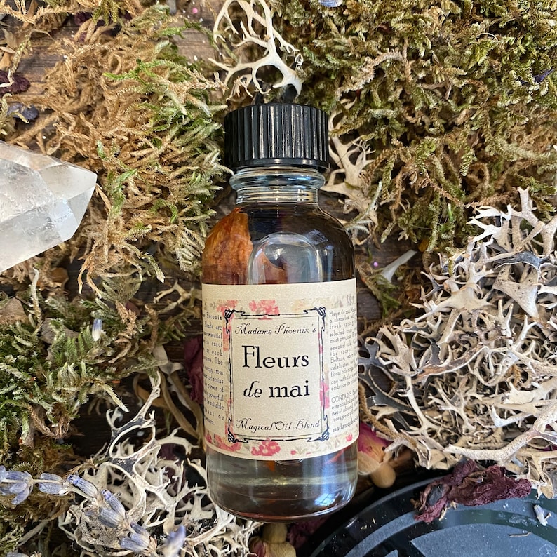 Może przedstawiać: Mała szklana butelka z czarną zakrętką wypełniona brązową cieczą. Etykieta zawiera napis "Madame Phoenix's Fleurs de mai Magical Oil Blend".