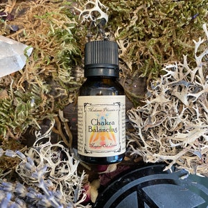 May include: A small brown glass bottle with a black cap, labeled "Madame Phoenix's Chakra Balancing Magical Oil Blend". The bottle is surrounded by moss and other natural elements.