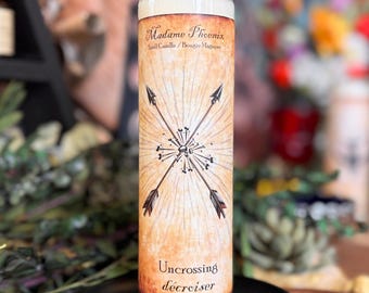 Könnte beinhalten: Eine hohe weiße Wachskerze in einem zylindrischen Glasbehälter. Das Etikett hat einen beigen Hintergrund im Pergamentstil mit dem Text Madame Phoenix, Spell Candle und Uncrossing. Eine Grafik mit zwei gekreuzten Pfeilen ist mittig platziert.