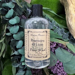May include: A clear bottle of Rosemary & Lime Floor Wash by Madame Phoenix's, with a black cap and a label featuring botanical illustrations. The bottle is surrounded by green and purple foliage.