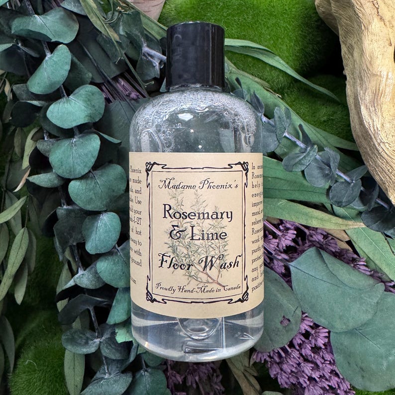 May include: A clear bottle of Rosemary & Lime Floor Wash by Madame Phoenix's, with a black cap and a label featuring botanical illustrations. The bottle is surrounded by green and purple foliage.