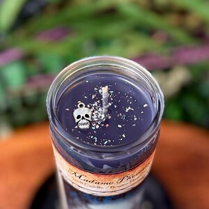 May include: A cylindrical glass candle filled with dark purple wax. The top surface features a central wick, scattered iridescent glitter, and a small silver-toned skull charm. A paper label on the glass reads Madame Phoenix Spell Candle.