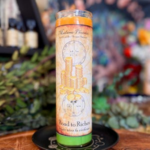 May include: A tall cylindrical glass candle with orange wax at the top and green wax at the bottom. The label features the text Madame Phoenix Spell Candle, an illustration of stacked gold coins, and the text Road to Riches with circular symbols.