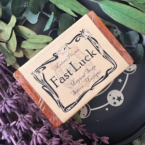 May include: An orange soap bar with a label featuring the text "Fast Luck Magical Soap Savon Magique" in black script. The label has a decorative black border. The soap is presented with dried lavender and green foliage.