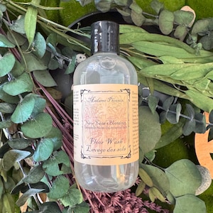 Könnte beinhalten: Eine klare Flasche Bodenreiniger mit schwarzem Verschluss. Auf dem Etikett steht "Madame Phoenix New Year's Blessing Floor Wash". Die Flasche ist von grünem Laub und lila Blumen umgeben.