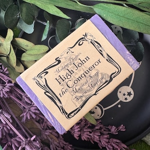 May include: A bar of soap with a lavender-colored wrapper and a tan label featuring the text "High John the Conqueror Magical Soap". The soap is surrounded by sprigs of lavender and green leaves on a black plate.