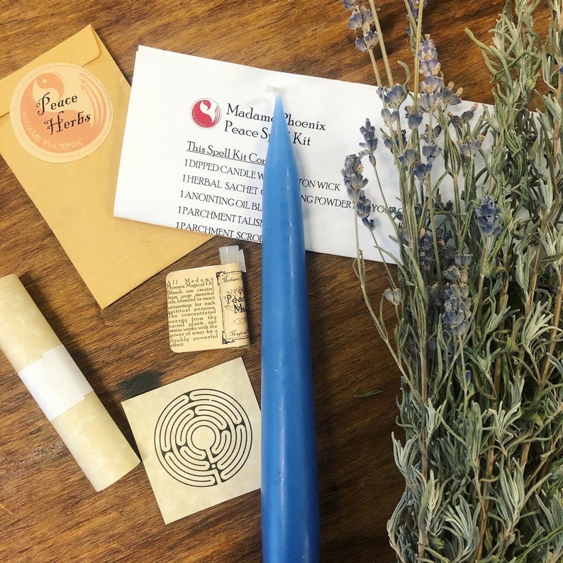 May include: A blue candle, a small bottle of oil, a rolled parchment, a small paper with a labyrinth design, a bundle of lavender, and a brown envelope with a sticker that says "Peace Herbs" are arranged on a wooden surface. The envelope also has the text "Madame Phoenix" on it. A white card with the text "Madame Phoenix Peace Spell Kit" is visible in the background.