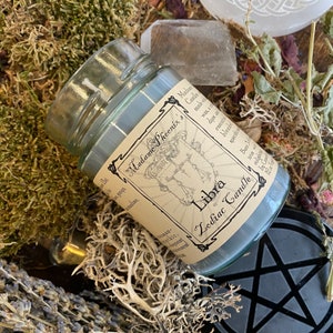 May include: A glass jar candle with a label that reads "Madame Phoenix's Libra Zodiac Candle". The label is decorated with a drawing of the Libra zodiac symbol and a scale.