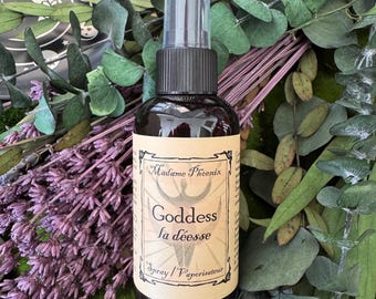 May include: A dark brown spray bottle with a black cap and clear top. The label reads "Madame Phoenix Goddess la déesse Spray / Vaporisateur". The bottle is surrounded by lavender and green foliage.