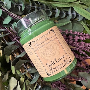 May include: A green candle in a glass jar with a label that reads "Madame Phoenix Spell Candle/Bougie Magique" and "Self Love Amour de Soi". The candle is surrounded by green and purple foliage.